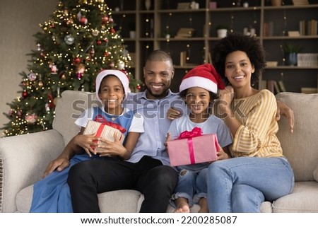 Happy Black family couple and sibling kids celebrating New Year at home. Young parents and children in Santa hats resting on sofa at Christmas tree, holding xmas gift boxes, smiling. Festive portrait