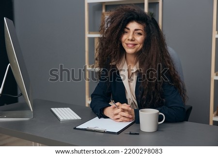 Cute happy friendly tanned adorable curly Latin businesswoman in jacket enjoy at work project smile at camera in office. Copy space Mockup Banner. Corporation leader lady uses modern computer in work