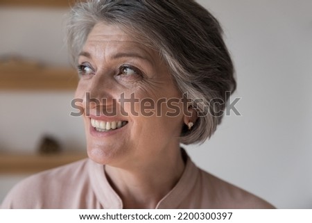 Happy dreamy retired grey haired woman looking away, with toothy smile, thinking, feeling joy. Face of cheerful thoughtful elder 60s lady close up portrait. Elderly age, retirement