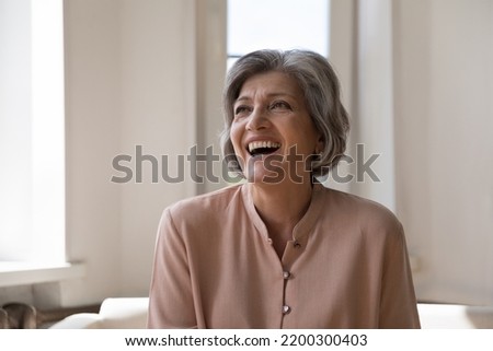Cheerful excited mature woman looking away, laughing, sitting on couch at home, feeling joy, rejoicing at good news, success. Grey haired 60s lady in casual getting happy