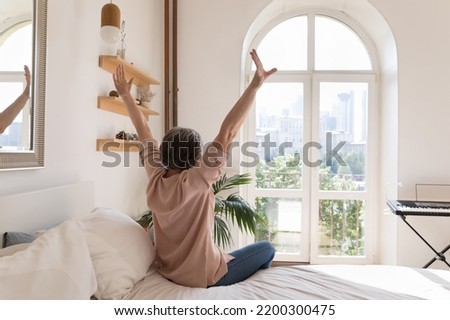 Relaxed older mature woman enjoying being in cozy home bedroom, good morning, sitting on bed, feeling great after sleeping enough on comfortable mattress, rising hands, stretching body. Back view Royalty-Free Stock Photo #2200300475