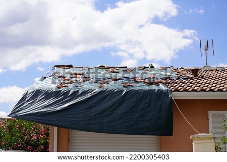 Storm damaged roof on house with a black plastic tarpaulin over hole in the shingles and rooftop after spring summer thunderstorm violent Royalty-Free Stock Photo #2200305403