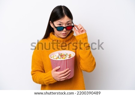 Young Chinese woman isolated on white background surprised with 3d glasses and holding a big bucket of popcorns