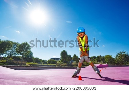 Young boy pose standing in blue helmet skate fast on rollerblades in the skatepark wearing protection and going around cones