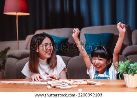 Asian happy family of little daughter play toy wooden block in home. Mother or mom young woman loving parent having fun play stack tower wood block game at night in living room. Hobbie leisure concept