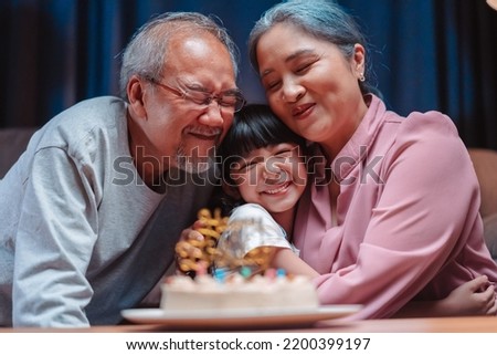 Asian happy family of little girl hugging lover with grandparents after blowing out candles on cake. Celebrate birthday anniversary party on table at night in living room, having happiness lifestyle. Royalty-Free Stock Photo #2200399197
