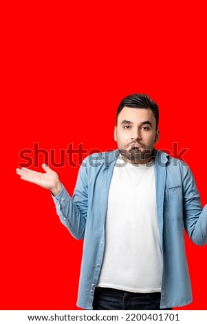 Good looking young man, looking surprised and unknowing, raises his shoulder and right hand Lake, isolated red background