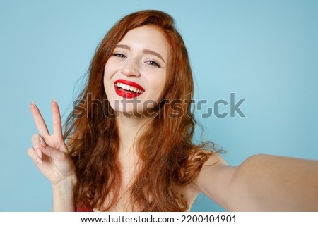 Close up young caucasian smiling redhead curly woman in red party evening dress gown doing selfie shot on mobile phone show victory v-sign gesture isolated on pastel blue background studio portrait