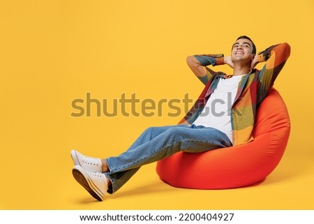 Full body young minded fun middle eastern man wear casual shirt white t-shirt sit in bag chair hold hands behind neck look overhead isolated on plain yellow background studio People lifestyle concept
