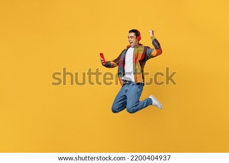 Full body young fun happy cool middle eastern man wears casual shirt white t-shirt headphones listen music jump high do winner gesture hold use mobile cell phone isolated on plain yellow background