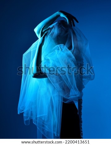 Close up portrait of beautiful woman model wearing elegant black dress and gauze shroud fabric, posing against a studio background with fantasy inspired arm gestures, multi coloured creative lighting.