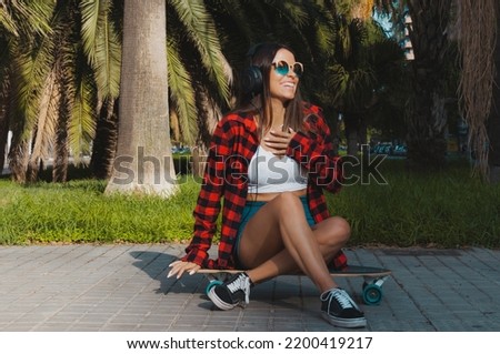 Laughing latin woman sitting on a skateboard listening to music and having fun, lifestyle.