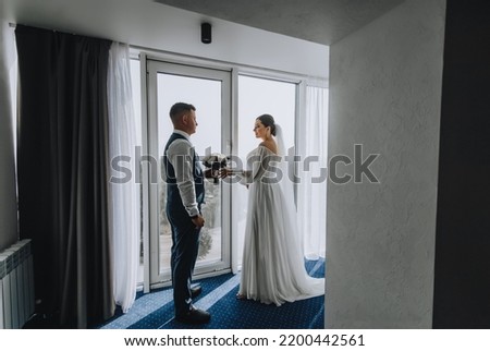 A stylish groom with a bouquet of roses and a beautiful bride in a white dress are standing near the window indoors, hotel. Wedding portrait, photography.