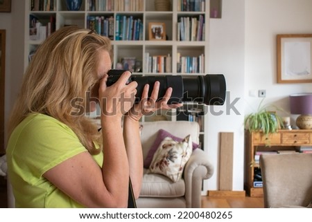 A woman takes a picture with a long lens DSLR camera. An attractive blonde lady looks into the viewfinder and composes a photograph. Sharp focus on the lady with soft focus on the colorful room behind