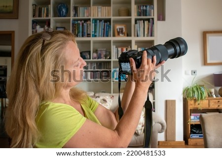 A woman adjusts a long lens DSLR camera. An attractive blonde lady looks at the back screen before she composes a photograph. sharp focus on the lady with soft focus on the colorful room behind