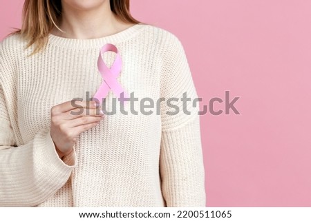 Female health check awareness. Closeup of woman hand in white pullover holding pink ribbon, symbol of breast cancer, timely diagnosis, support of oncology patients. indoor isolated on pink background