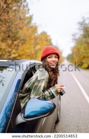 Beautiful woman takes pictures on the camerafrom the car window. Smiling woman enjoying autumn weather. Rest, relaxation, lifestyle concept.