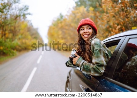 Beautiful woman takes pictures on the camera from the car window. Smiling woman enjoying autumn weather. Rest, relaxation, lifestyle concept.