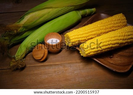 Ripe maize cob in green leaves and boiled corn on the cob in the wooden plate with saltcellar on teh wooden table. Still-life corn abstract picture