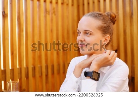 Portrait of calm smiling woman with bun hairstyle wearing white shirt, sitting with palms together and looking away, dreaming, being in good mood, resting.
