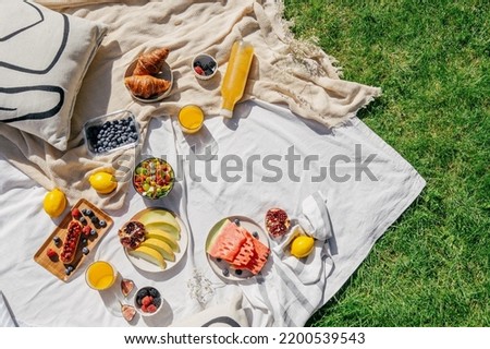 top view of white tablecloth with different food, juicy fruits, ripe berry, vegetables salad in bowl, sweet croissants, fresh orange juice in glass near bottle, plaid and pillow on grass outdoors Royalty-Free Stock Photo #2200539543