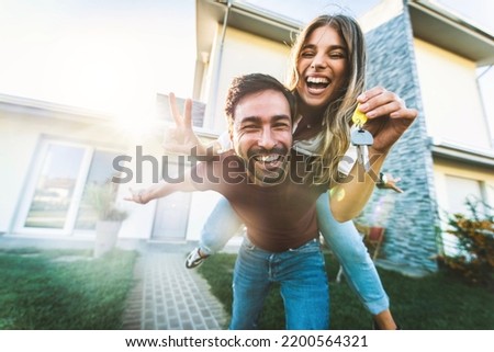 Happy young couple holding home keys after buying real estate - Husband and wife standing outside in front of their new house