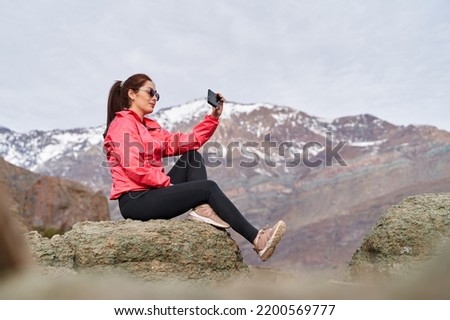 young red haired woman with her hair taken, taking pictures with her phone, wearing a red jacket, sitting on a rock in the middle of the Andes Mountains of Chile