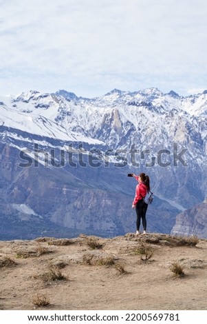 young red haired woman with red jacket and backpack, taking pictures with her phone in the middle of the Andes mountain range in Chile