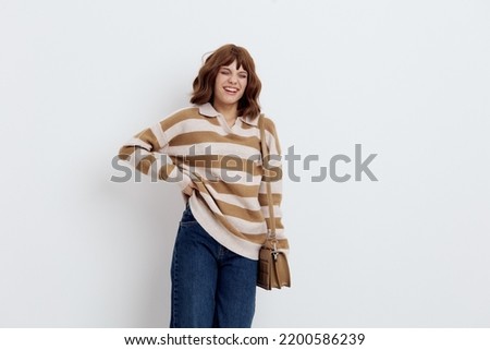 a joyful woman stands on a white background in a striped sweater and with a bag on her shoulder put her hand on the belt pulling the sweater up a little, putting her other hand on the bag