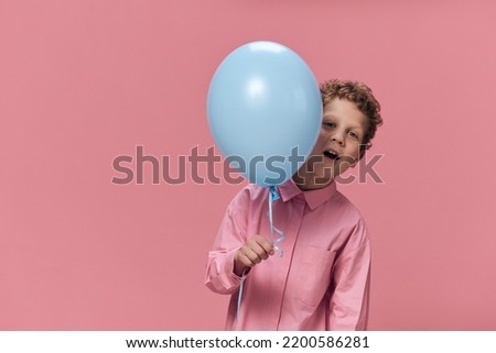 a cute, funny boy of school age stands on a pink background in pink clothes and funny hides his face behind a big blue balloon in his hand.