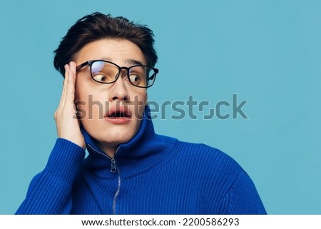 a handsome, attractive man stands on a light blue background in a blue zip-up sweater and holding black-rimmed glasses on his face with his hand, looks in surprise at the camera with his mouth open