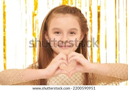 Kid's fashion and beauty. Pretty girl in a golden evening dress poses on a white background with shiny gold foil curtains showing a heart sign and smiling.  Close up shot.