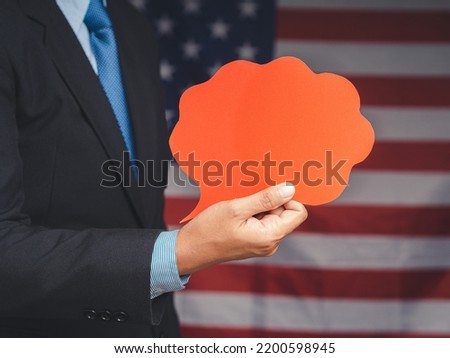 Speech bubble concept. Close-up of a hand businessman in a suit holding a blank red speech bubble while standing on the American flag background. Space for text