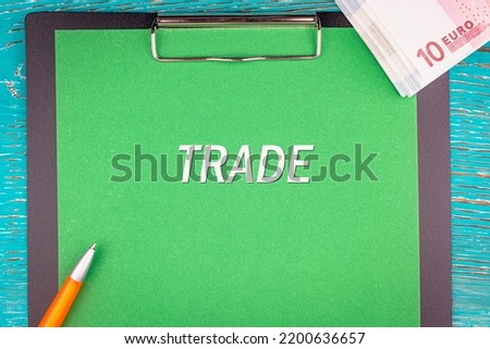 TRADE - word (text) and a bundle of money, euro bills on a green background of a notebook, a pen and a wooden table. Business concept: buy, sell, commerce (copy space).