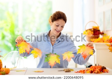 Autumn home decoration. Family decorating house with colorful maple leaves banner. Thanksgiving or Halloween celebration in white living room. Woman with fall leaf. Creative crafts ideas with foliage.