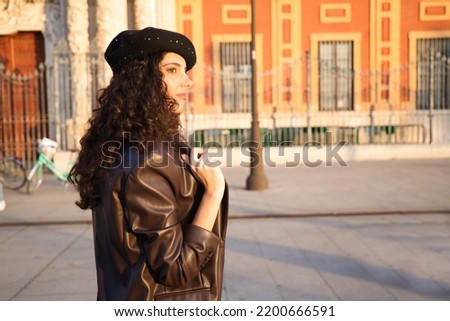 Pretty young woman with curly brown hair wearing a black cap and dressed in a leather jacket, jeans and white boots. She is sightseeing in the city. Concept of holidays and tourism around the world. Royalty-Free Stock Photo #2200666591
