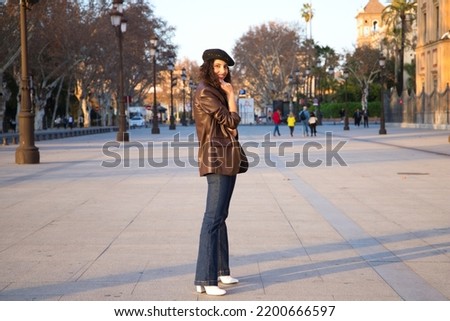 Pretty young woman with curly brown hair wearing a black cap and dressed in a leather jacket, jeans and white boots. She is sightseeing in the city. Concept of holidays and tourism around the world.