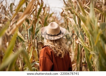 Stylish woman with red coat and cowboy hat is standing in corn field. Autumn season in farm