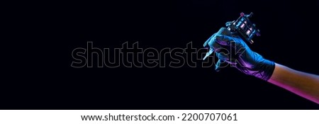 Closeup of tattooer master's hand in black glove holding machine for making tattoo art on body isolated on dark background in neon. Concept of art, job, hobby, professional equipment. Flyer for ad Royalty-Free Stock Photo #2200707061