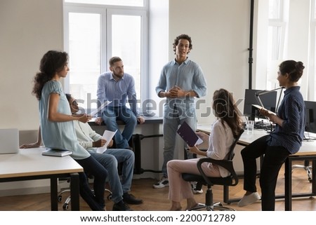 Young Latin leader guy presenting idea for startup project to diverse group on business team meeting, talking to colleagues. Corporate mentor, coach giving consultation to employees Royalty-Free Stock Photo #2200712789