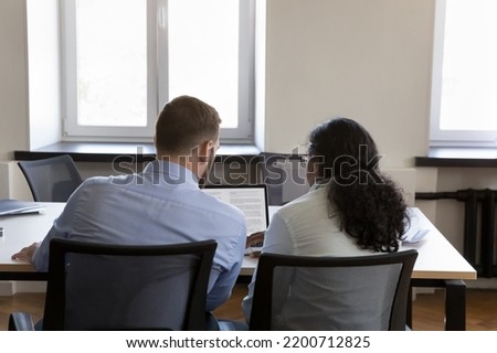 Two multiethnic employee men sharing laptop at table, looking at display. Mentor and intern reading, editing text together, working on business article. Corporate education, teamwork concept