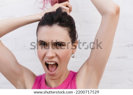 Portrait of young pink hair woman in pink top, plays with hair, arms raised up, photo on light brick background.  Girl make high ponytail and screaming. Happy attractive woman braids her hair. 