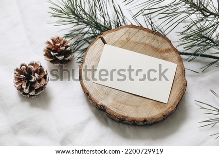 Christmas stationery still life. Blank business card, invitation mockup on cut wooden round board. Pine cones and green Christmas tree branches on white linen cloth background. Top view, no people.