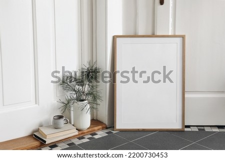 Minimal festive Christmas indoor decor. Blank vertical wooden picture frame mockup on tiled floor. Pine tree branches in vase, cup of coffee and old books. White hall background. Empty template. 