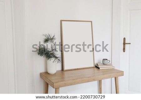 Christmas Scandinavian interior. Blank vertical wooden picture frame mockup. Pine tree branches in vase, cup of coffee and old books on table, desk. White wall background, doors. Empty copy space. 