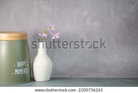 White ceramic vase with purple flower, green-metal jar with text 'Happy Moment' on its. Bright and clean background for your editing. 