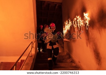 Brave Fireman Descends Stairs of a Burning Building and Holds Saved Girl in His Arms. Open fire and one Firefighter in the Background.
