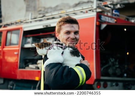 Close-up portrait of heroic fireman in protective suit and red helmet holds saved cat in his arms. Firefighter in fire fighting operation.