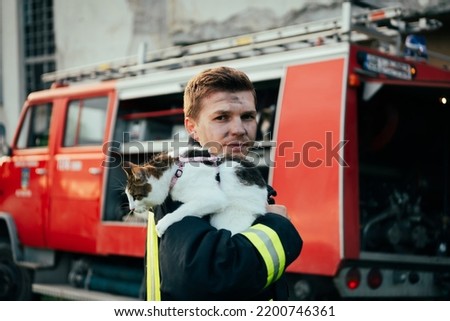 Close-up portrait of heroic fireman in protective suit and red helmet holds saved cat in his arms. Firefighter in fire fighting operation. Royalty-Free Stock Photo #2200746361