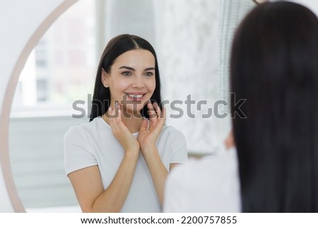 Portrait of a woman looking in the mirror at home, touching her face, skin, smiling, admiring Royalty-Free Stock Photo #2200757855